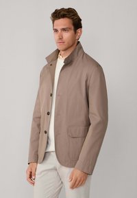 Veste légère couleur beige avec col montant, boutonnage devant, poche poitrine unique et texture lisse, associée à un pull crème.