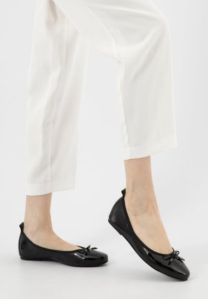 Frau trägt weiße Hose und schwarze Ballerinas aus Lackleder mit kleinem Schleifendetail, steht auf einem Fuß.