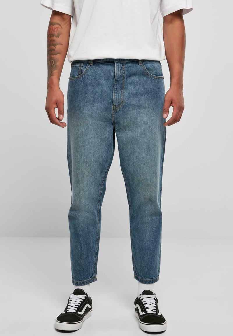 Hombre con vaqueros azules desgastados, calcetines blancos, zapatillas negras y blancas, y una camisa blanca de manga corta, con antebrazos tatuados visibles.