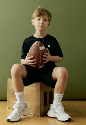 Mlajši fant v črnih športnih oblačilih sedi na lesenem zaboju, drži rjavo ameriško nogometno žogo, nosi bele nogavice in superge, ob zeleni steni.