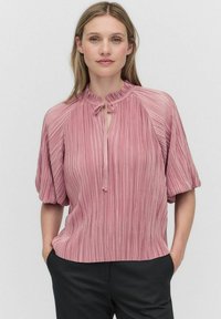 Vaaleanpunainen laskostettu pusero, jossa on röyhelökaulus, silmukkatyyppinen yksityiskohta ja lyhyet puffihihat. Kangas näyttää pehmeältä, ja siinä on pystysuorat raidat.