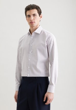 Homme portant une chemise blanche à boutons avec de fines rayures verticales violettes et un pantalon plissé bleu marine foncé, debout devant un fond uni.