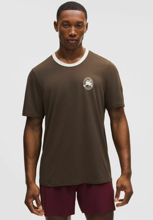 Mand iført mørkebrun kortærmet skjorte med hvid krave og lille hvid grafik på brystet, kombineret med vinrøde shorts, stående mod ensfarvet baggrund.