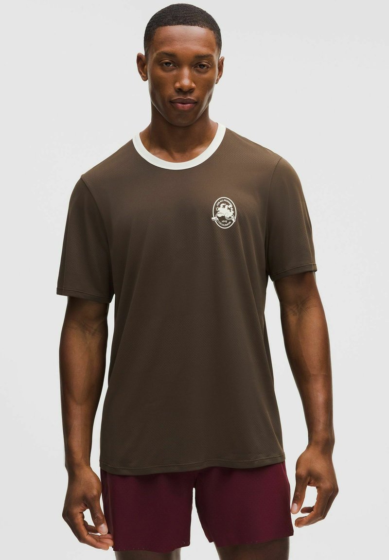 Férfi sötétbarna rövid ujjú ingben, fehér gallérral és a mellkason kis fehér grafikával, bordó rövidnadrággal, unalmas háttér előtt állva.