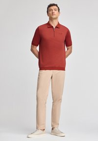 Röd pikétröja med dragkedja, korta ärmar och ribbad nederkant; tillsammans med beiga byxor och ljusa sneakers. Enkelt design och slät textur.