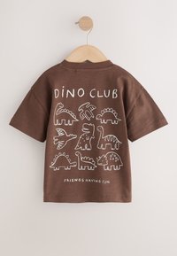 Camiseta marrón de manga corta con dibujos blancos de dinosaurios y el texto "Dino Club" y "Friends Having Fun" colgada en una percha de madera.