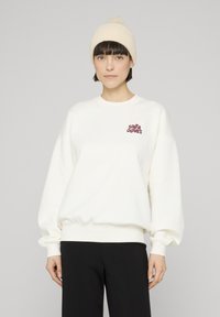 Krēmkrāsas džemperis ar ribveida aprocēm un apakšmalu, ar bordo krāsas logotipu. Saskaņots ar melnām biksēm un vieglu cepurīti.