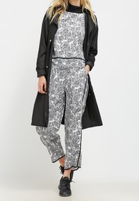 Un long manteau noir sur un top et un pantalon à motif floral monochrome, offrant une coupe décontractée et un parement contrastant. Des baskets noires complètent l'ensemble.