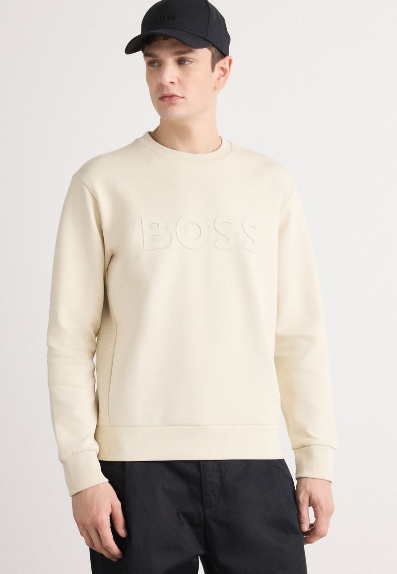Heledbeige sweatshirts, millepakkunud "BOSS" logo, ümmargune kaelus ja pikad varrukad. Kandmise ajal must kork ja tumedad püksid.