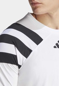 Hvid atletisk skjorte med sort-hvide striber på skuldrene, ribbet krave og synligt Adidas-logo i sort. Glat tekstur.