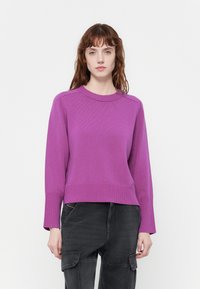 Pull en maille côtelée violet, à col rond et à manches longues. Porté avec un pantalon cargo noir ample.