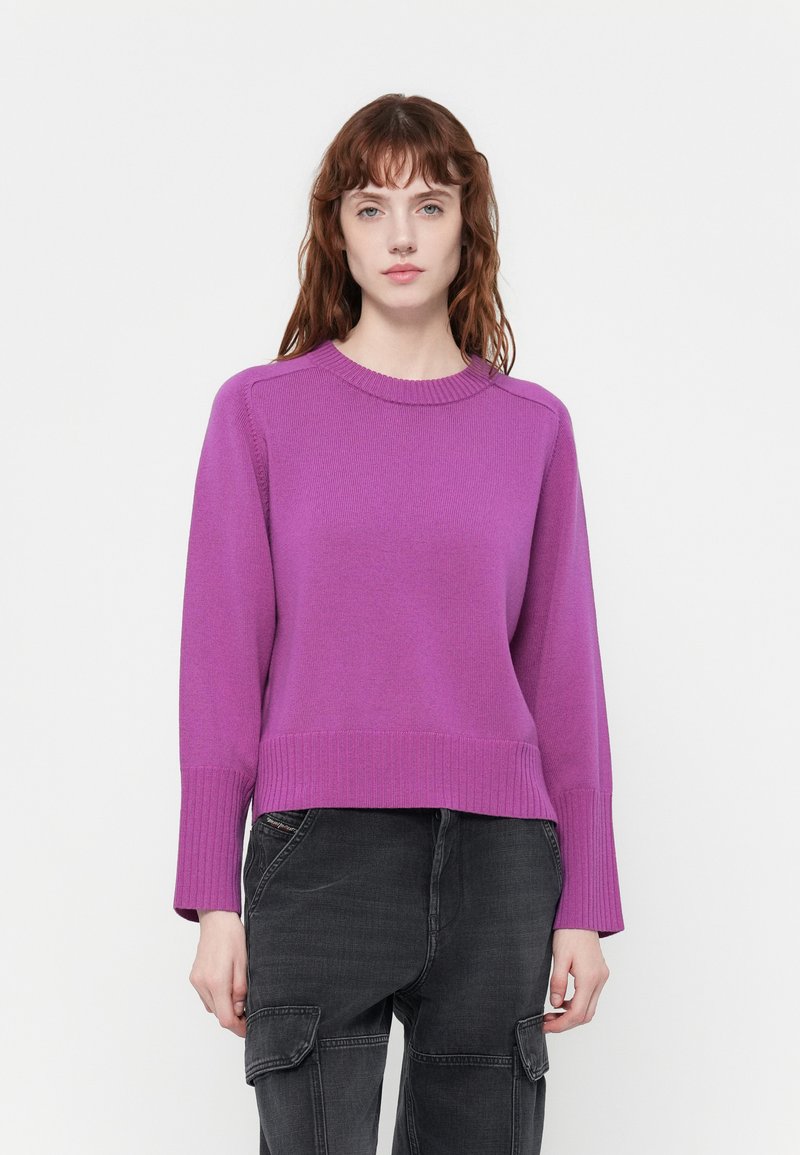 Lila, gerippter Strickpullover mit Rundhalsausschnitt und langen Ärmeln. Kombiniert mit weiten, schwarzen Cargo-Hosen.