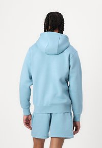 Lyseblå hettegenser med snor, ribbet mansjetter og hem, laget av mykt stoff. Kombinert med matchende shorts, begge har en jevn tekstur.