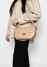 Borsa a tracolla in pelle rosa con design strutturato, dettaglio della chiusura dorata e tracolla, portata da una persona con una camicia in finta pelle color crema.