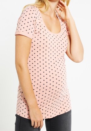 Femme portant une chemise rose à manches courtes avec des pois noirs et un pantalon foncé, touchant son cou avec une main.