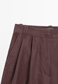 Pantalon plissé marron taille haute avec passants pour ceinture et poches latérales, fabriqué en tissu texturé.