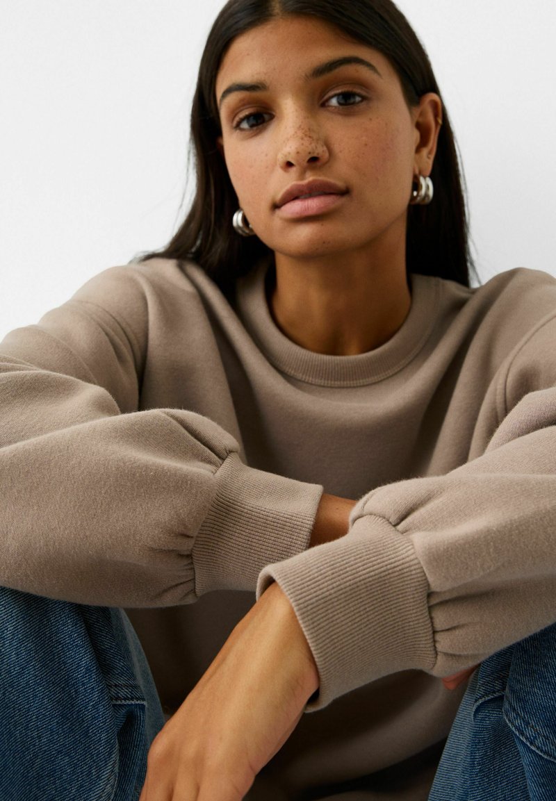 Felpa beige con collo a girocollo a coste e maniche a palloncino oversize, abbinata a jeans blu. Tessuto morbido e vestibilità rilassata.