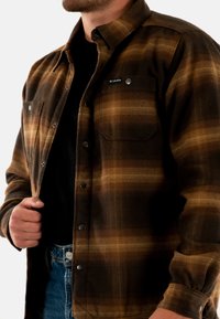 Brązowa i pomarańczowa koszula flanelowa w kratę, z guzikiem z przodu, dwiema kieszeniami na piersiach i subtelnymi akcentami metalowymi. Posiada miękką teksturę.