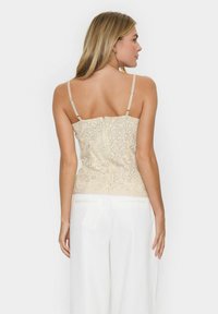 Top in pizzo di colore beige chiaro con spalline sottili, busto aderente e scollo a cuore. Motivo floreale testurizzato sul tessuto.