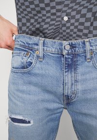 Personne portant un jean bleu clair déchiré et une chemise à carreaux noire et grise, tenant la taille du jean.