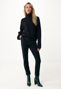 Suéter negro de punto con cuello alto combinado con pantalones negros ajustados y botas de tacón verde. Texturas suaves, diseño ajustado y acentos sutiles.