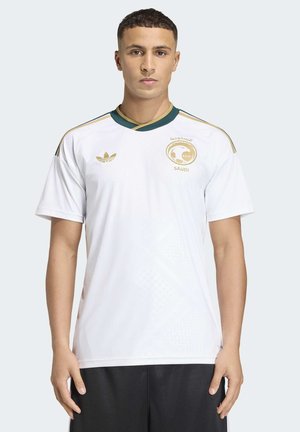 Junger Mann trägt weißes saudi-arabisches Fußballtrikot mit goldenen und grünen Details, steht vor hellem einfarbigem Hintergrund.