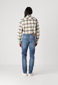 Kostkovaná flanelová košile v béžové, modré a hnědé barvě, která se nosí s lehkými modrými džínami, vybavenými koženou nášivkou a standardními zadními kapsami.