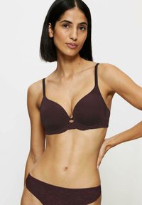 Brązowy stanik z formowanymi miseczkami i regulowanymi ramiączkami, ozdobiony koronkowym pasem i centralnym wycięciem. Widoczny jest również dopasowany brązowy dół bikini.