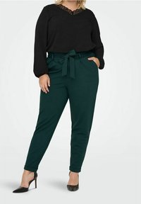 Woman wearing black long-sleeve top with lace neckline, dark green high-waisted trousers with belt, and black pointed heels, standing.