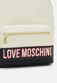 Sac à dos en toile avec une partie supérieure crème, un bas noir et un texte brodé "LOVE MOSCHINO" en rouge. Quincaillerie en doré sur la poche avant.