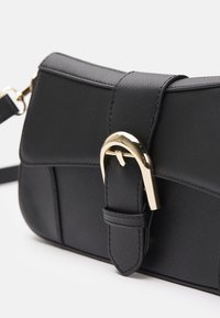 Sac d'épaule en cuir noir avec surface texturée et fermeture à boucle incurvée dorée sur le rabat avant, présenté sur fond blanc.