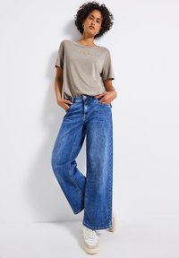 Camiseta de manga corta gris claro con el texto "puro y seguro" combinada con unos vaqueros de pierna ancha de mezclilla azul. Zapatillas blancas con acentos en beige.