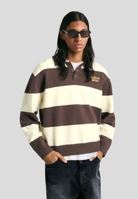 Hosszú hajú személy fekete napszemüvegben, barna és krémszínű csíkos pulóverben gombokkal és sötét nadrágban, kezeit a zsebében tartva áll.