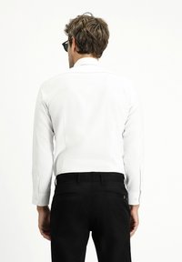 Camicia bianca a maniche lunghe con polsini a bottoni, abbinata a pantaloni neri. La camicia ha una texture liscia e un colletto standard.
