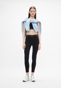 Čierne priliehavé legíny s logom Calvin Klein, čierny skrátený top a svetlo modrý sveter prehodený cez plecia; v kombinácii s bielymi teniskami.