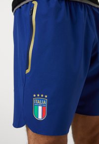 Calções desportivos azuis, feitos de um tecido leve, com um bolso com fecho dourado e um logótipo bordado da FIGC Itália com quatro estrelas acima.