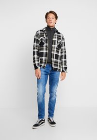 Rutet flanellskjorte i svart og hvitt med gule detaljer, over en grå turtleneck, kombinert med blå jeans og svarte joggesko.