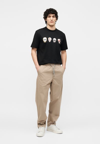Νέος άνδρας στέκεται με τα χέρια στις τσέπες, φοράει μαύρο μπλουζάκι με τέσσερα πολύχρωμα κρανία, μπεζ παντελόνι και λευκά αθλητικά παπούτσια.