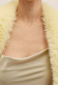 Zbliżenie na osobę w beżowej satynowej sukience z asymetrycznym dekoltem oraz puszystą, jasnoniebieską szalą z futra.