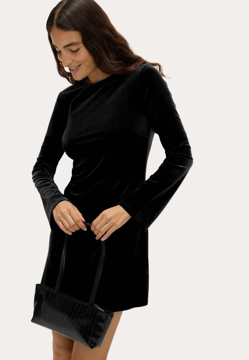 Schwarzes Samtkleid mit langen Ärmeln, kombiniert mit einer strukturierten schwarzen Handtasche. Das Kleid hat einen Rundhalsausschnitt und die Handtasche hat eine rechteckige Form.