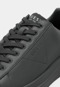 Baskets Guess noires avec des lacets épais, logo embossé sur le côté et texte imprimé sur la languette indiquant "GUESS Est. 1981."