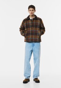 Kockovaná vlnená bunda v hnedých, modrých a oranžových odtieňoch, s golierom a bočnými vreckami, kombinovaná so svetlomodrými džínsami a hnedými čižmami.