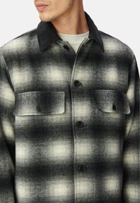 Giacca a camicia in tartan bianco e nero realizzata in tessuto testurizzato con colletto in corduroy nero, dotata di due tasche sul petto e bottoni neri.