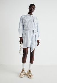 Svītrainas krekla kleitas gaiši zilā un baltā krāsā, ar pogu aizdari priekšā, divām krūšu kabatām, jostu viduklī un garām piedurknēm. Valkā ar bēšiem sporta apaviem.