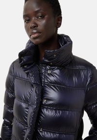 Granatowa kurtka puchowa z wysokim kołnierzem, błyszczącą fakturą i złotymi zatrzaskami. Posiada poziome przeszycia i ukrytą zamek błyskawiczny.