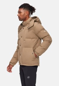 Giacca puffer color cammello con cappuccio, dotata di chiusura a snap, tasche laterali e tessuto testurizzato. Indossata su pantaloni neri con logo.