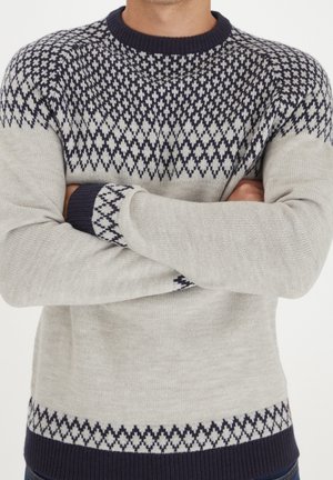 Persona con un maglione grigio con un motivo a rombi blu navy sul petto, sui polsini e sull'orlo, con le braccia incrociate sul torso.