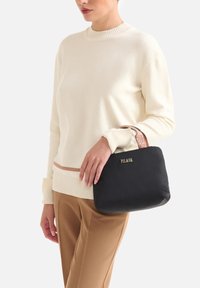 Borsa nera con manico testurizzato, logo dorato e materiale morbido. La modella indossa un maglione color crema con un sottile accento a righe e pantaloni color cognac.