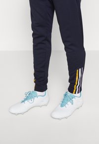 Botas de fútbol blancas con una superficie texturizada y acentos en azul claro, combinadas con pantalones navy que presentan rayas coloridas en los tobillos.
