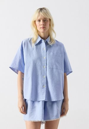 Blonde woman wearing a loose light blue button-up shirt and matching shorts with white lace trim, standing against a plain white background.